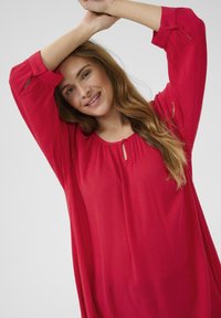 Blouse rouge ample à manches longues et col rond, avec un petit détail en forme de clé. Fabriquée en tissu léger et texturé.
