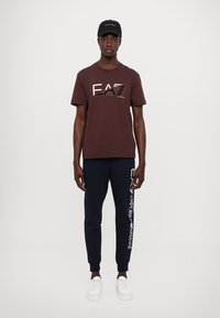 Barna pamut póló "EA7" logóval, tengerészkék melegítőnadrág oldalsó márkajellel, fehér sportcipő és fekete sapka, modell frontális nézetben, fehér háttér előtt.