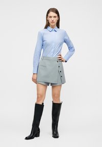 Γυναίκα που στέκεται με το χέρι στη μέση, φοράει ανοιχτό μπλε ριγέ πουκάμισο, γκρι ασύμμετρη φούστα-σορτς και μαύρες μπότες μέχρι το γόνατο.