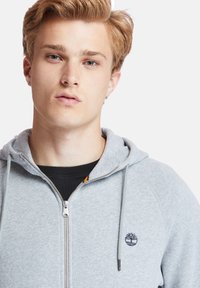 Felpa con zip di colore grigio chiaro realizzata in misto cotone. Presenta una zip frontale, un cappuccio con cordini e un piccolo logo sul petto. Camicia nera sotto.