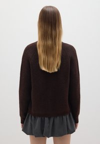 Suéter marrón de punto con un patrón texturizado, puños acanalados y un corte relajado, llevado sobre una falda gris con pliegues, visto desde la parte de atrás.