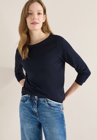 Femme aux cheveux châtain clair portant une chemise à manches longues bleu marine et un jean en denim bleu sur un fond uni clair.