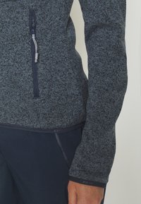 Texturerad, mörkt grå fleece-pullover med en dragkedjeficka, kontrasterande fållar och åtsittande manschetter. Mjuk tyg med ett fläckat mönster.