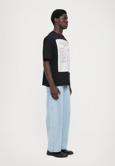 T-shirt en coton noir à coupe oversized, avec un imprimé graphique blanc sur le devant. Une paire de jeans évasés bleu clair et des chaussures noires.
