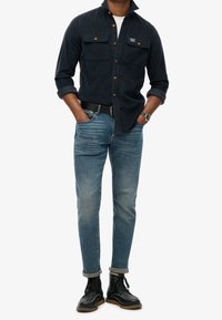 Granatowa koszula z kordurowego materiału z dwiema kieszeniami na piersiach i brązowymi guzikami, zestawiona z niebieskimi zwężającymi się dżinsami i czarnymi botkami na sznurówki.