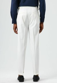Pantalon blanc sur mesure avec coupe droite, ourlets retroussés et deux poches arrière, associé à des chaussures noires et une chemise bleu marine.