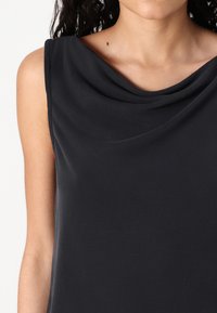 Haut à col montant en tissu gris foncé, présentant une texture lisse et un design sans manches, accentué par un drapage délicat autour de l'encolure.