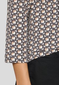Ärmel eines geometrisch gemusterten Oberteils in Beige, Weiß und Dunkelbraun, getragen mit schwarzen Hosen, die Nähte und Stofftextur zeigen.