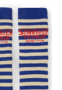 Calzini a righe blu e crema con la scritta "Stranger Things Benetton" in rosso vicino al polsino superiore su uno sfondo bianco.
