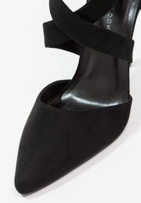 Chaussure en daim noir à bout pointu avec des brides croisées sur une semelle intérieure noire brillante, sur fond blanc.