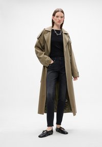 Khaki színű trench coat, fekete ing, sötét farmernadrág és fekete cipő. A kabát laza fazonú, nagy hajtókákkal és részletes fém kiegészítőkkel rendelkezik.