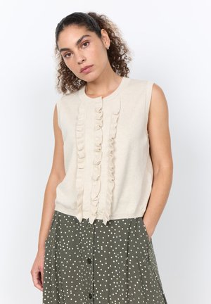 Frau mit lockigem Haar, die eine ärmellose cremefarbene Rüschenbluse und einen hoch taillierten olivgrünen Rock mit weißen Punkten und Knöpfen trägt.
