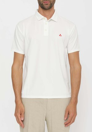 Mann trägt ein schlichtes weißes kurzärmeliges Poloshirt mit kleinem rotem Logo auf der linken Brustseite und beigen Hosen.