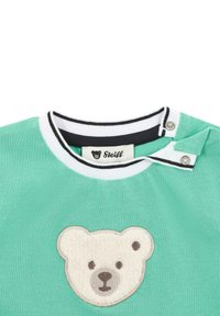 Camiseta de algodón verde con un apliqué de cara de oso texturizado. Presenta un cuello de rayas en blanco y negro y un cierre a presión en el escote.