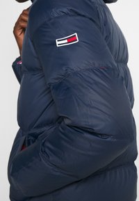 Námořnická puffer bunda s lesklým povrchem, s logem na rukávu, horizontálním švem a bočními kapsami orámovanými červeně.