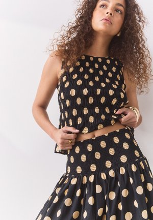 Femme aux cheveux bouclés porte un haut noir sans manches et une jupe à gros pois beiges, tenant l'ourlet de son haut.