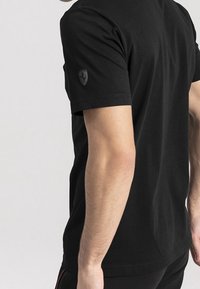 Camiseta de algodón negra de manga corta, con un pequeño logo gris en la manga izquierda. Textura suave y corte clásico.