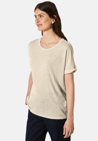 Beige kortärmad t-shirt tillverkad av ett mjukt tyg; har rund hals och rullade ärmslut. Lätt lös passform och sprund på sidorna.