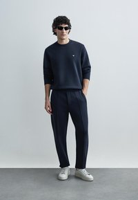 Tmavomodrá mikina s okrúhlym výstrihom a nenápadným logom, v kombinácii s tmavomodrými plisovanými nohavicami a bielymi teniskami. Model má na sebe slnečné okuliare.