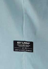 Tela de color azul claro con textura acanalada, que presenta una etiqueta negra con la inscripción "KEY LARGO", indicando el nombre de la marca y el eslogan en texto blanco.