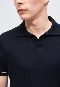 Νέος άνδρας που φοράει μαύρο πικέ πόλο με σχιστή γιακά και λευκή ρίγα στο μανίκι, μπροστά σε απλό φόντο.