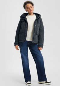 Zwarte geïsoleerde jas met een capuchon, voorzien van voorzakken en een ritssluiting, gecombineerd met een crème trui en lichtblauwe wijde jeans.