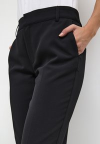 Black tailored trousers with a smooth texture, featuring side pockets and a slim fit, finished with a standard waist and belt loops.