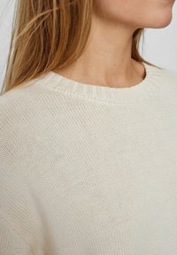 Maglione lavorato a maglia beige chiaro con un colletto rotondo a coste e un tessuto testurizzato. La vista ravvicinata mette in evidenza il motivo a maglia e la morbidezza del tessuto.