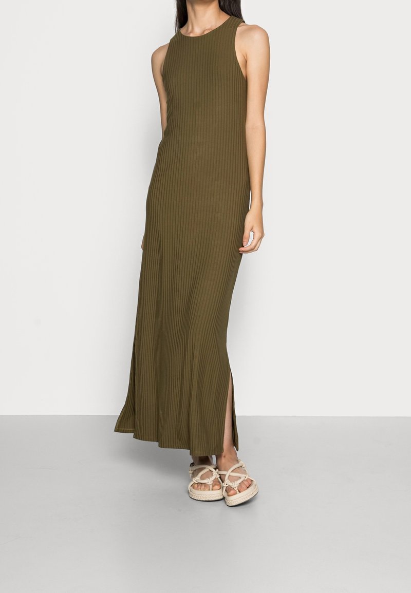 Robe maxi côtelée vert olive avec un col rond, des fentes latérales et une coupe ajustée, associée à des sandales beige à lanières.