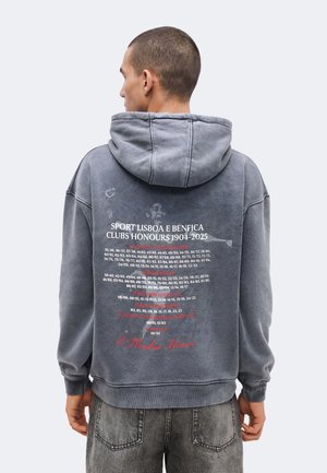 Osoba oblečená v sivej mikine s kapucňou, ktorá na chrbte vymenúva ocenenia klubu Sport Lisboa e Benfica od roku 1904 do 2025.