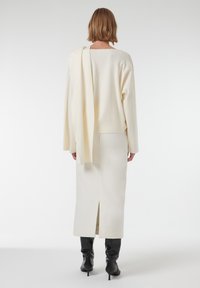 Crème trui over de schouder gedrapeerd, gecombineerd met een crèmekleurige midi-rok met een split aan de achterkant. Model draagt zwarte hoge hakken.