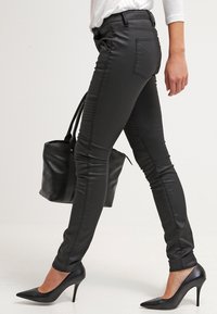 Pantalones skinny de imitación cuero negros con una textura suave, diseño ajustado y acentos acanalados en la parte frontal. Combinados con tacones altos negros y un bolso.