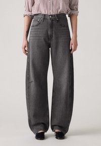 Persona valkāja augsto jostasvietu, brīvu, izbalējušu melnu bikses, melnus apavus un sarkanu ar baltu vertikāli svītrotu krekli ar uzceltām piedurknēm.