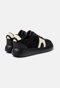 Svarta sneakers med ett texturerat tygöverdrag och beige accenter, som har en tjock svart sula och ett bakre dragband för enkel påtagning.