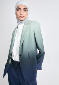 Ombre-Blazer, der von hellgrün nach dunkelblau übergeht, mit strukturierter Passform, Reverskragen und einem einzelnen schwarzen Knopf. Getragen über einem weißen Oberteil.