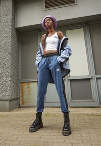 Modré tepláky s černými bočními pruhy, bíl�é crop top, a nadměrná modrá bunda. Obsahuje fialovou čepici a masivní černé boty.