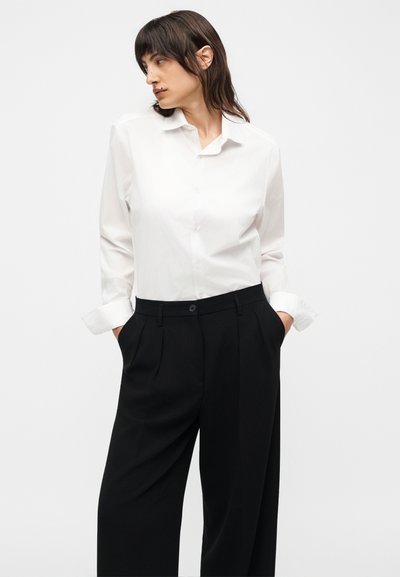 Άτομο με κοντά σκούρα μαλλιά που φοράει λευκό πουκάμισο με κουμπιά και μαύρο παντελόνι ψηλόμεσο με φαρδιά πόδια, τα χέρια στις τσέπες, κοιτάζει προς τα αριστερά.