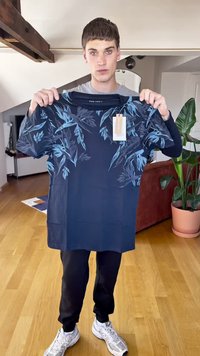 Mornarsko modra majica s kratkimi rokavi z svetlo modrimi listnatimi vzorci, iz bombažnega materiala, z vidno etiketo. Model drži majico v prostoru z lesenim tlakom.