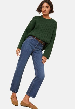 Grüner Strickpullover mit strukturiertem Muster, blaue gerade Jeans und brauner Gürtel, kombiniert mit beigen flachen Schuhen.