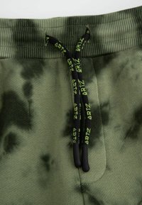 Zielone spodnie z tie-dye z prążkowanym pasem w talii, czarnym sznurkiem z zielonymi akcentami oraz miękkim, teksturowanym materiałem.