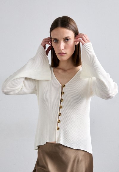 Λευκό ριπ φούτερ με V-ντεκολτέ, διακοσμημένο με επτά χρυσά κουμπιά μπροστά και φαρδιές καμπάνες στα μανίκια, συνδυασμένο με ένα μεταξένιο καφέ φούστα.