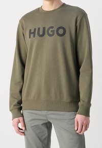 Olivově zelená mikina se kulatým výstřihem a elastickými manžetami. Velké černé logo "HUGO" přes hrudník, vyrobené z hladké látky.