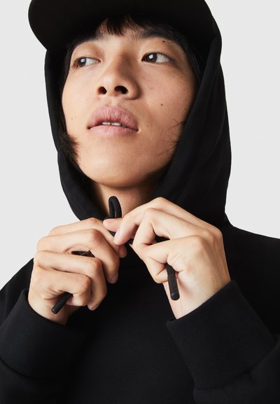 Sweat-shirt noir avec cordon de serrage, porté avec des mains ajustant la zone du cou, mettant en avant une texture douce et un design simple.