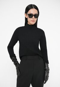 Czarny dzianinowy golf z teksturowanym wzorem w połączeniu z czarnymi spodniami z wysokim stanem. Modelka ma na sobie czarne skórzane rękawiczki i okulary przeciwsłoneczne.