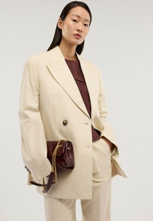 Frau in übergroßem cremefarbenem Anzug und bordeauxrotem Oberteil, die eine bordeauxrote Ledertasche mit goldener Kette vor hellem Hintergrund hält.