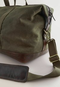 Grøn kanvas duffel taske med bund af brunt læder, stofhåndtag og en aftagelig skulderrem. Har lynlåslukning og messinghardware.