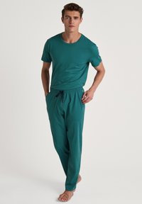 Teal t-shirt en bijpassende joggingbroek van zacht materiaal, met een elastische tailleband en twee zijzakken, staand model, blote voeten.