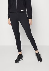 Persona vestida con leggings negros, chaqueta negra con cremallera y zapatillas Nike negras, frente a un fondo de suelo y pared blanca.