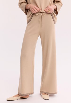 Osoba nosząca beżowe szerokie spodnie z sznurkiem w talii, pasujący top z paskowanym obszyciem i beżowe płaskie buty.