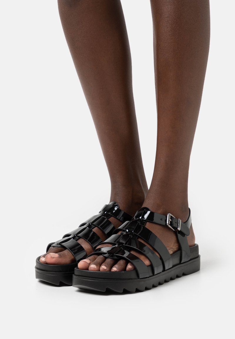 Jambes à la peau foncée portant des sandales gladiateur en cuir verni noir avec des boucles et des semelles épaisses et crantées sur un fond clair uni.
